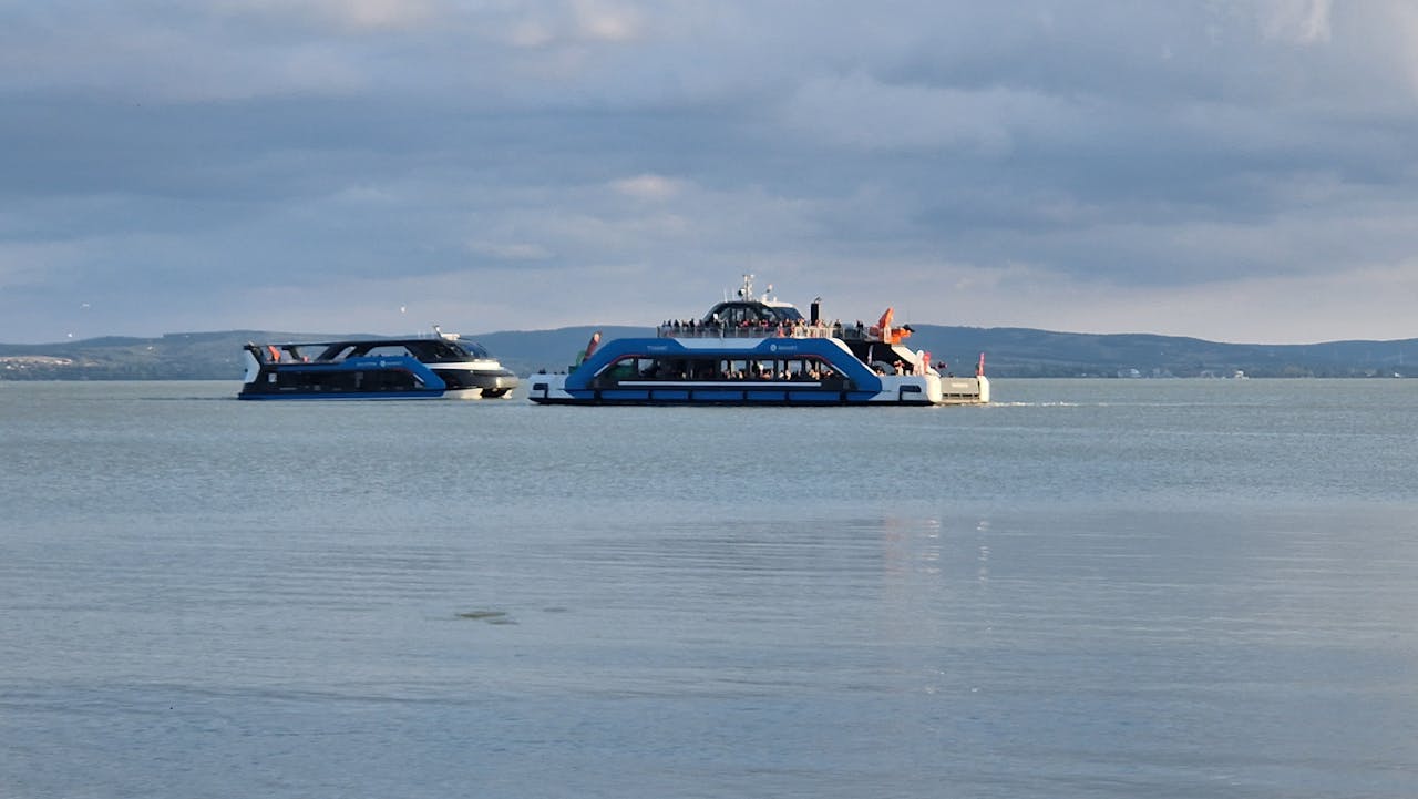 Two passenger ferries navigate a tranquil lake with hills in the background on a cloudy day.